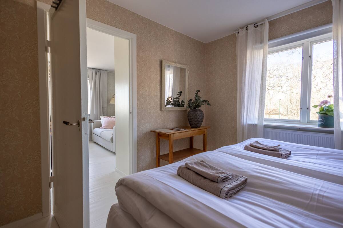 Een slaapkamer met twee bedden, een nachtkastje met een vaas, een raam met gordijnen en een deur die naar een andere kamer leidt.