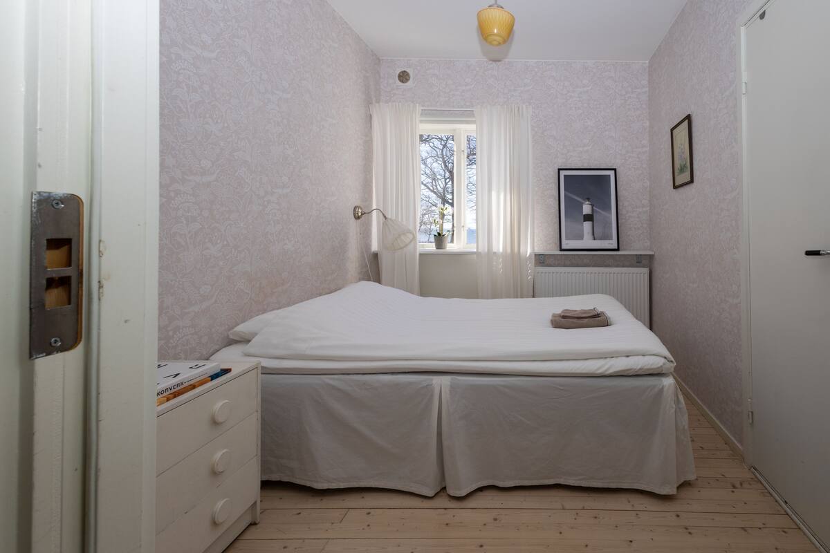 Een slaapkamer met een bed, een dressoir, een raam met gordijnen, een radiator en een ingelijste foto aan de muur.