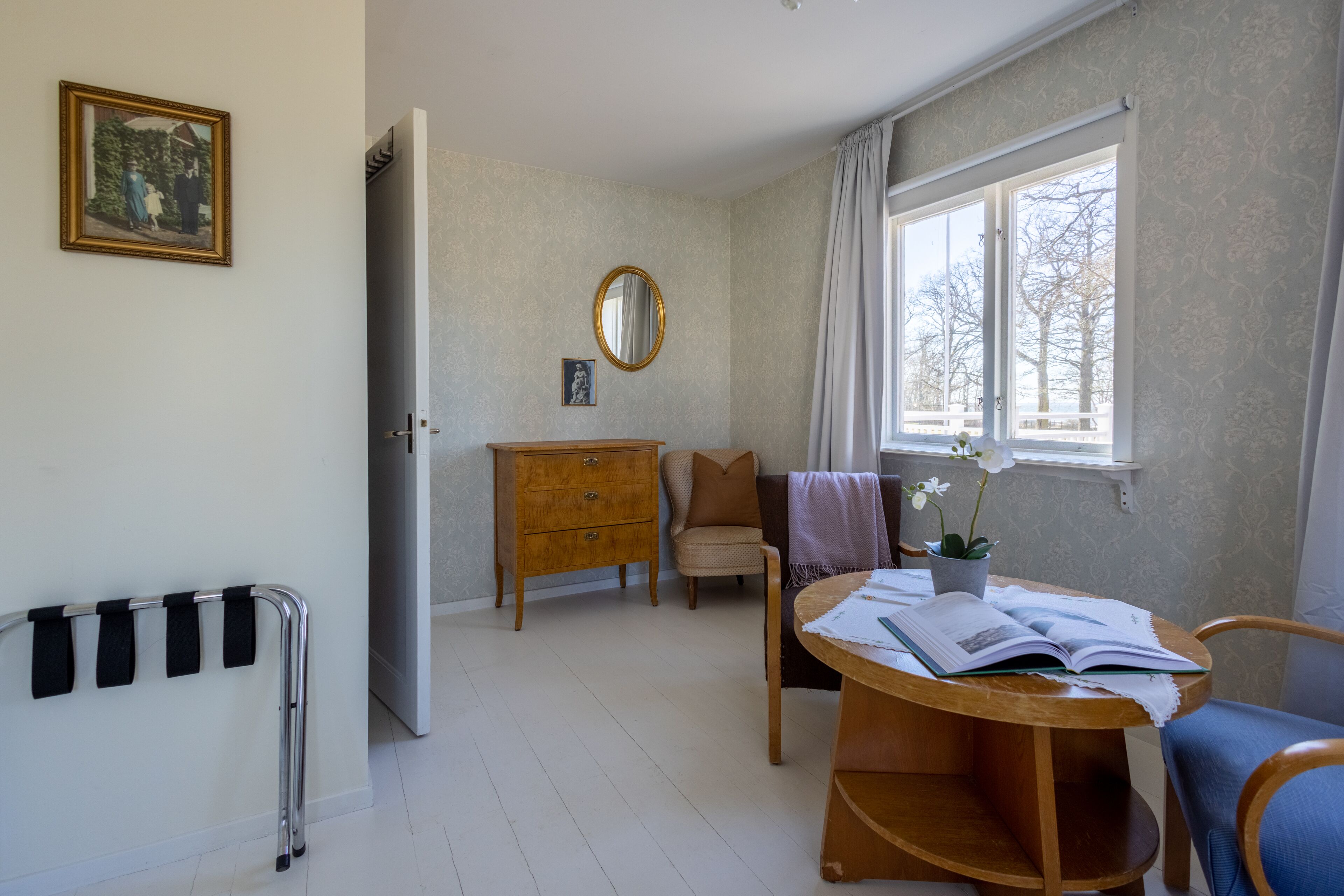 A room with a wooden table, a chair, a small cabinet, a mirror, and a window with curtains.