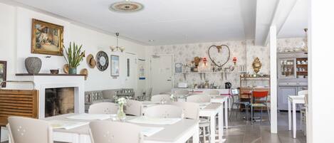 A dining area with white tables and chairs, a fireplace, and a kitchen in the background.