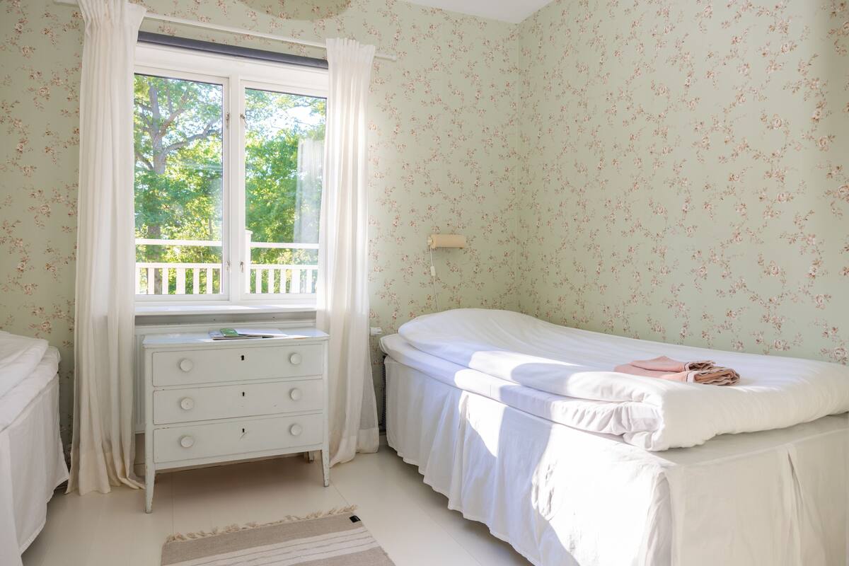 Een slaapkamer met een enkel bed, een nachtkastje, een dressoir en een raam met uitzicht op bomen.