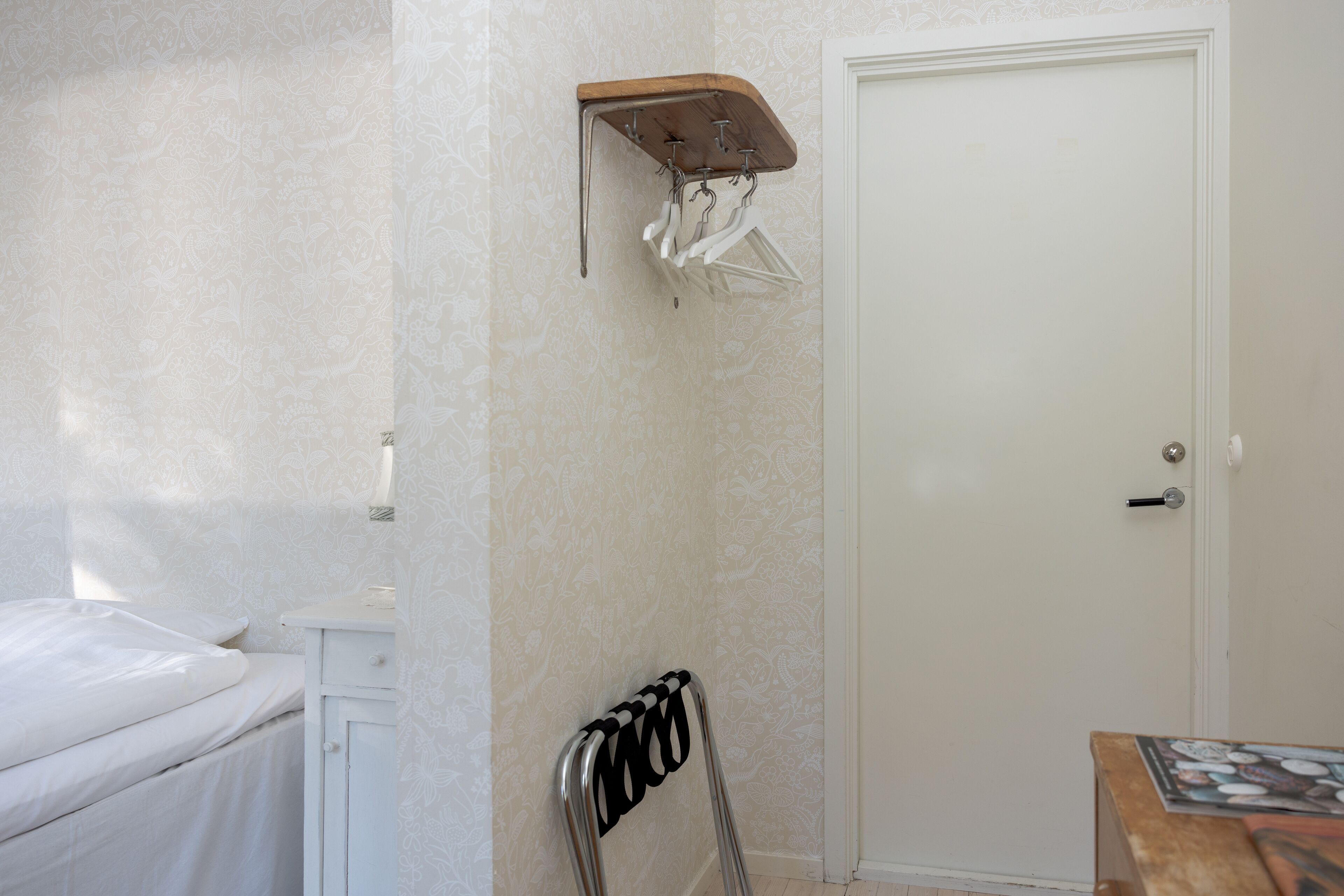 A bedroom with a bed, a white wardrobe, a wooden shelf with clothes, a metal drying rack, and a door.