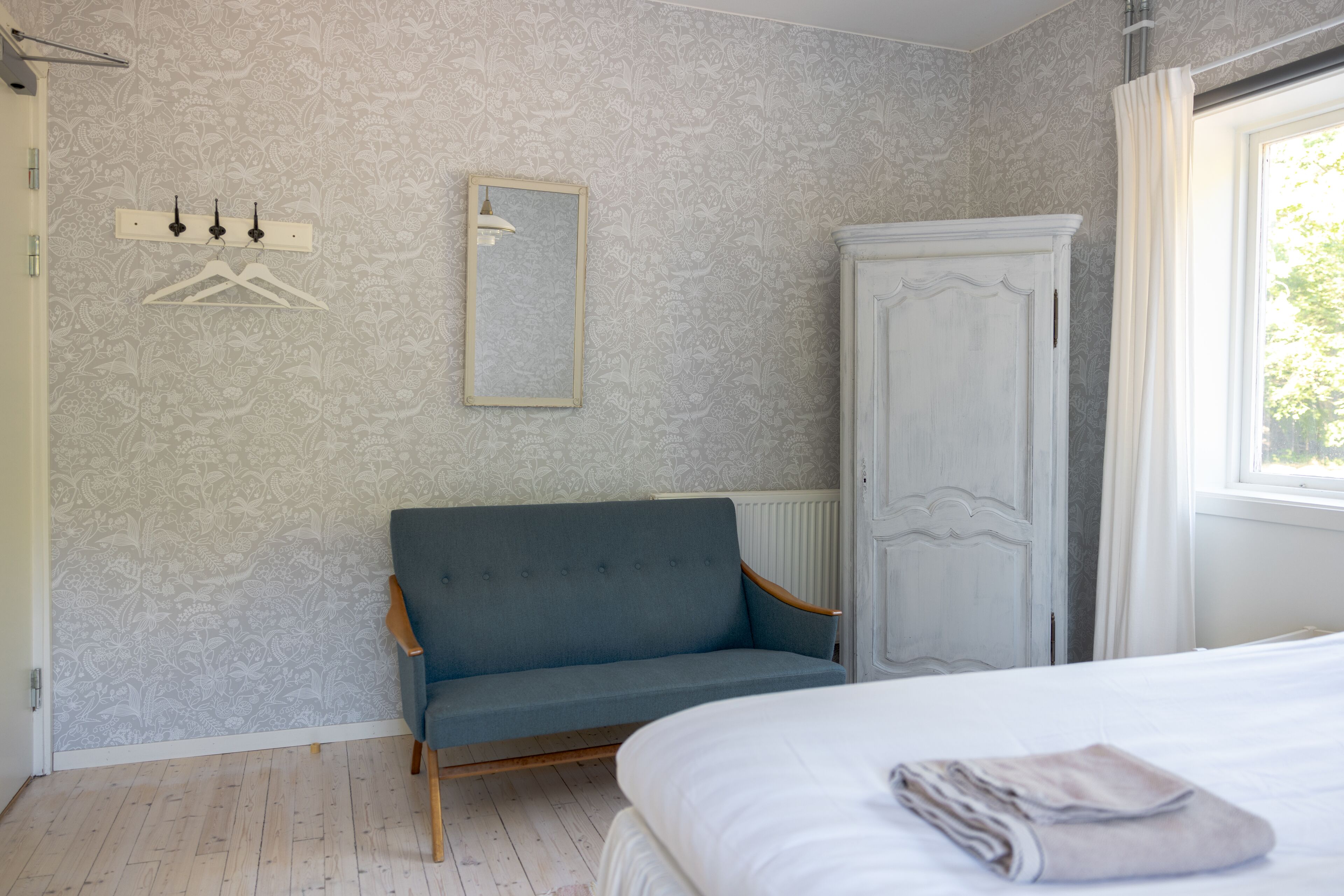 Un dormitorio con cama, una silla, un armario, un radiador y una ventana con cortinas.