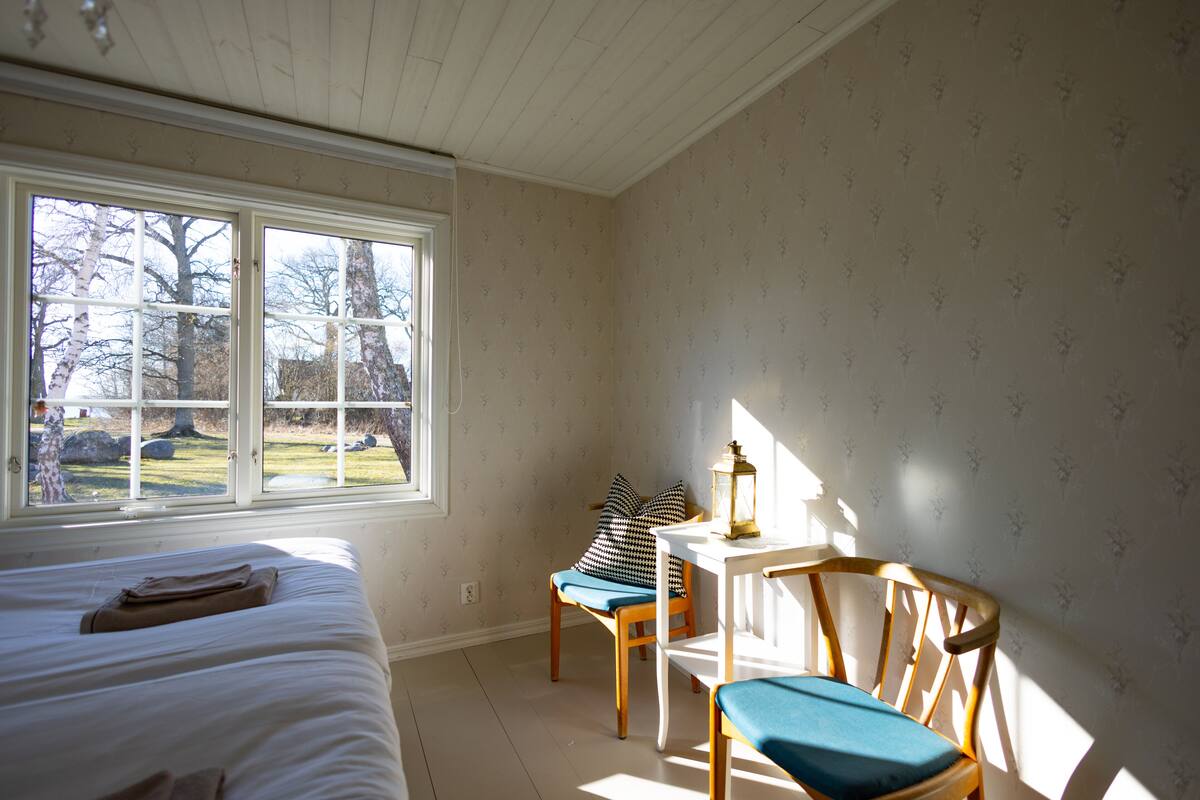 Een slaapkamer met een bed, een raam, een stoel en een tafel met een lamp.