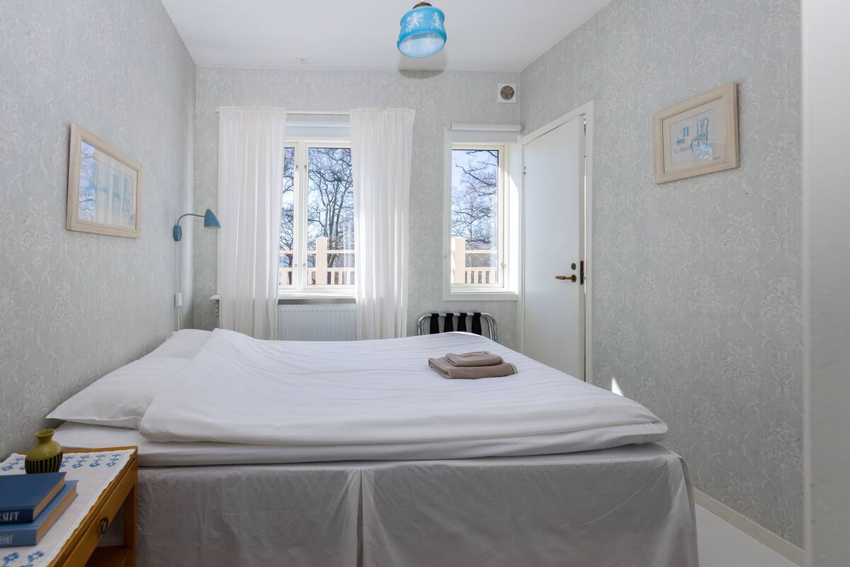 Een slaapkamer met een bed, een nachtkastje met boeken, een lamp en een raam met gordijnen.