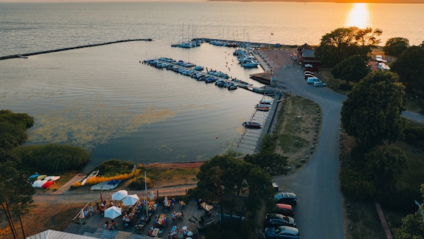 An aerial view of a marina with boats docked, a dock extending into the water, and a road leading to a building with outdoor seating. - Pensionat Strandhagen (Farjestaden)