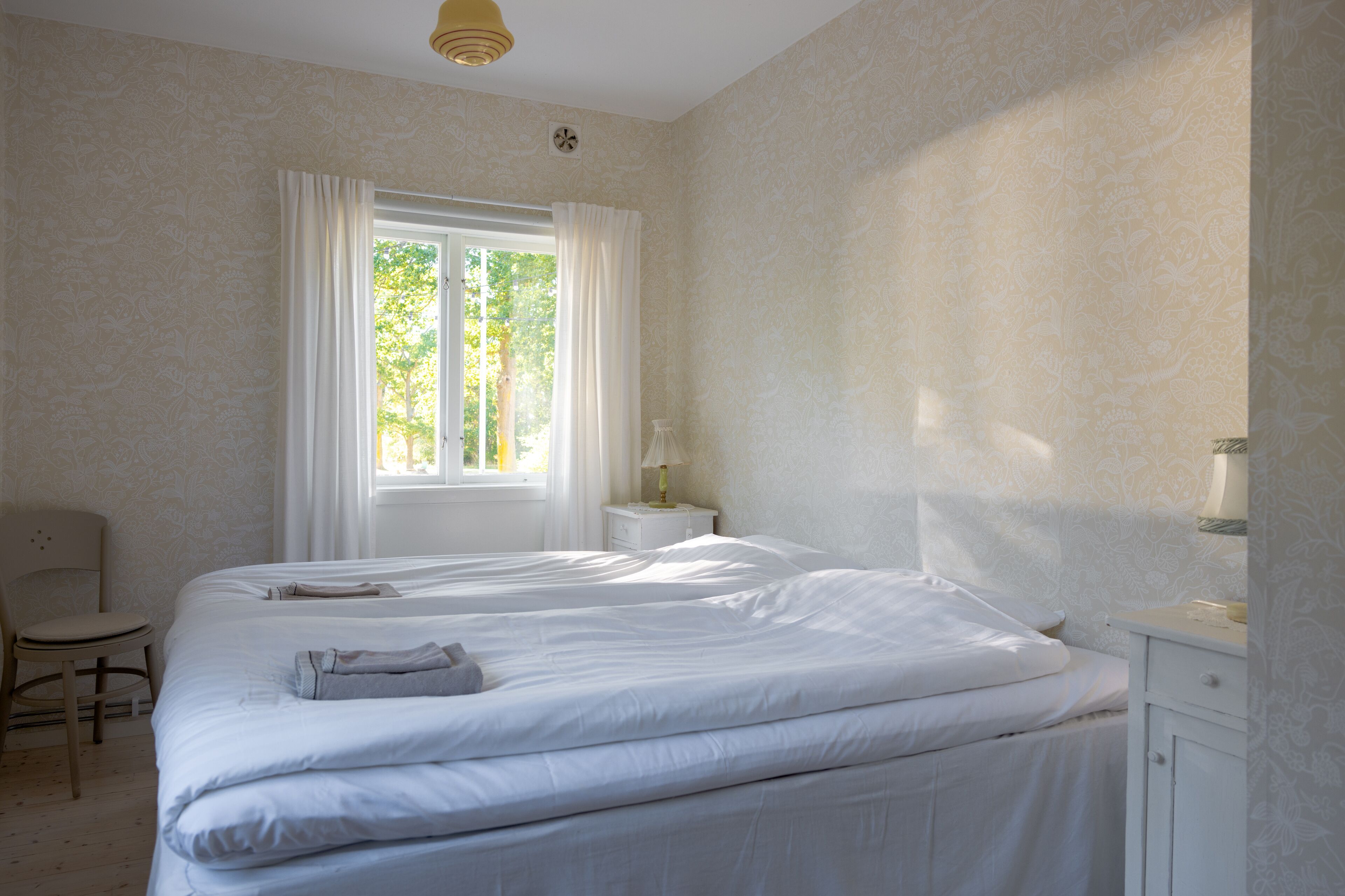 A bedroom with two beds, a chair, a nightstand, and a window with curtains.