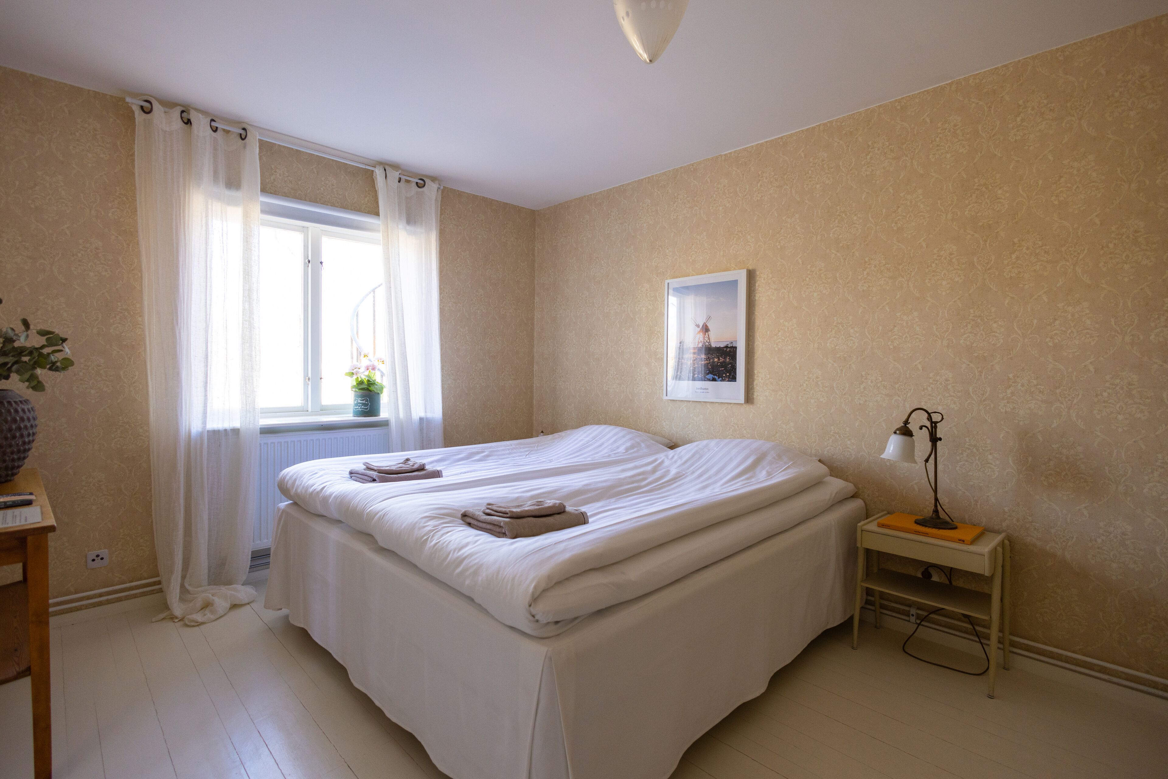 A bedroom with two beds, a window with curtains, a bedside table with a lamp, and a framed picture on the wall.