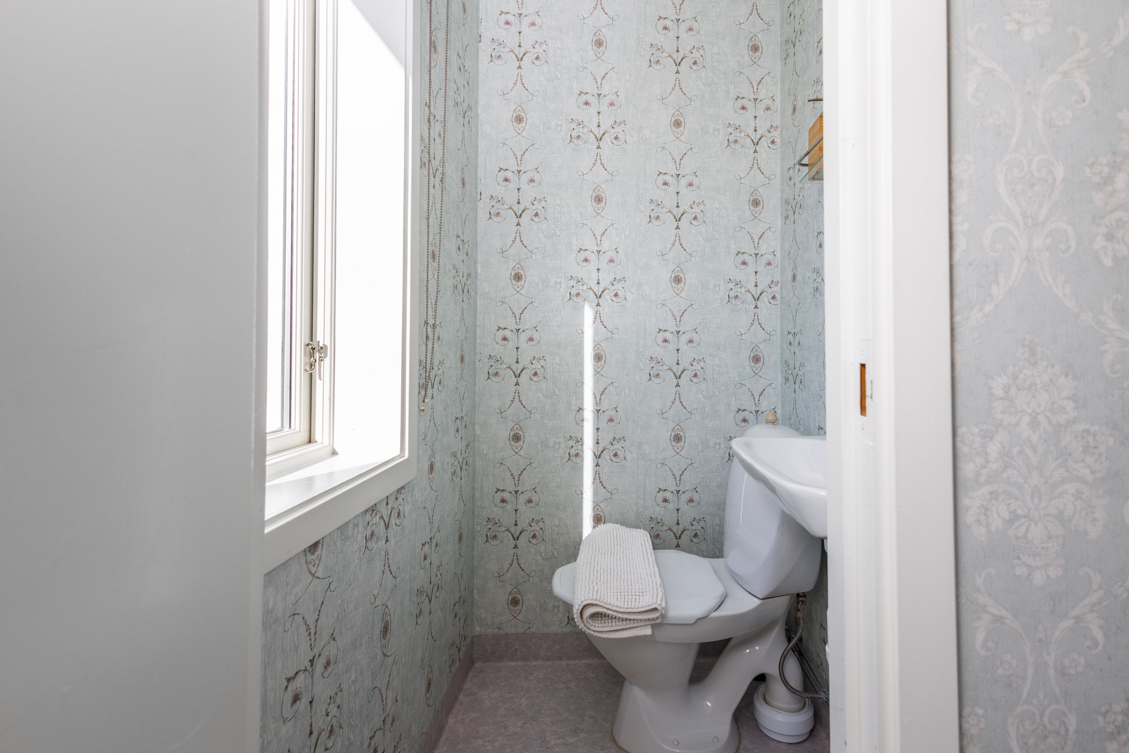 A bathroom with a toilet, a window, and patterned wallpaper.
