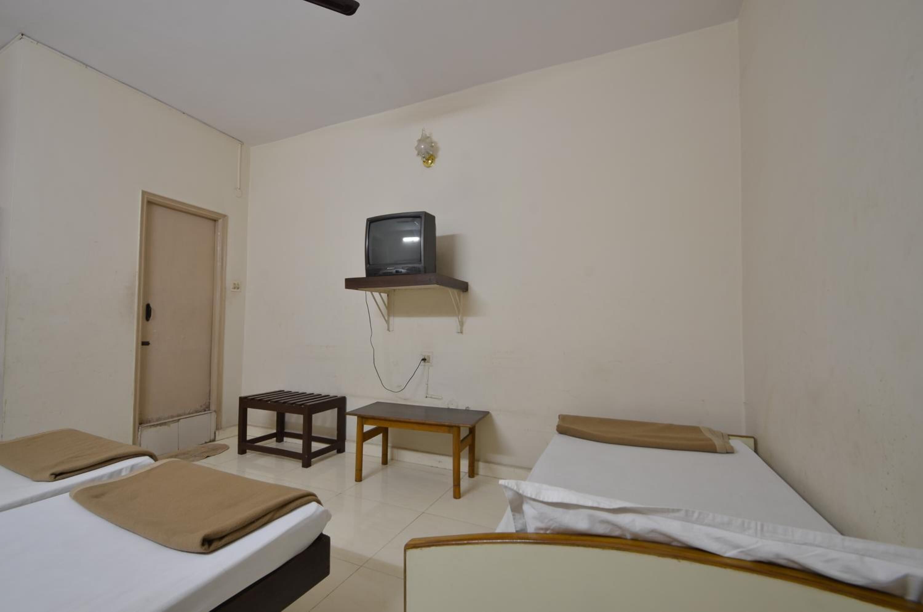 Ein kleines, schlichtes Zimmer mit zwei Betten, einem kleinen Tisch, einem Fernseher auf einem Regal und einer Tür.