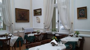 Un coin repas avec des tables dressées pour quatre personnes, chacune recouverte d’une nappe verte et agrémentée de chemins de table blancs. Deux tableaux ornent les murs, un vase rempli de fleurs est posé sur le rebord de la fenêtre, et une statue se trouve sur la cheminée.