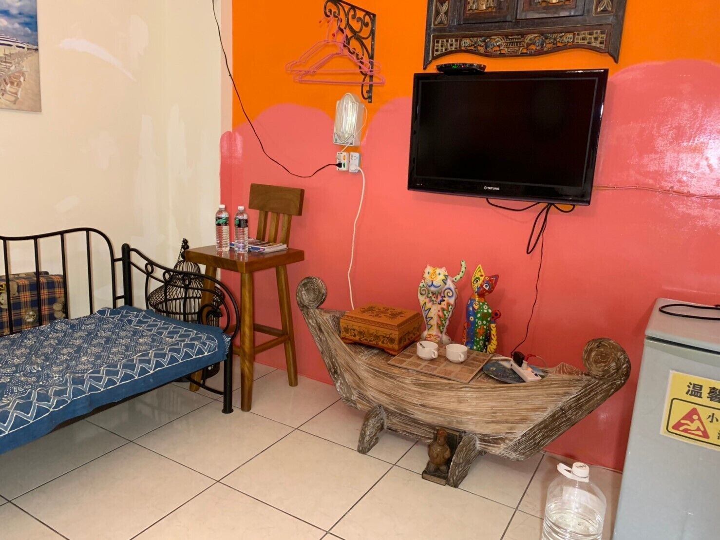 Una habitación pequeña con cama, una silla, una mesa con objetos decorativos, un televisor y un dispensador de agua.