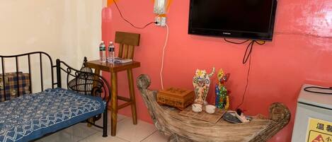 Una habitación pequeña con cama, una silla, una mesa con objetos decorativos, un televisor y un dispensador de agua.