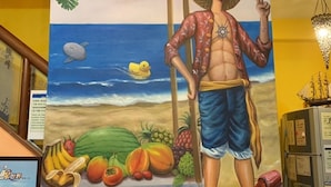 Un mural colorido con una persona de pie en la playa, sosteniendo un sombrero de paja, con una exhibición de frutas tropicales y una tortuga marina al fondo.