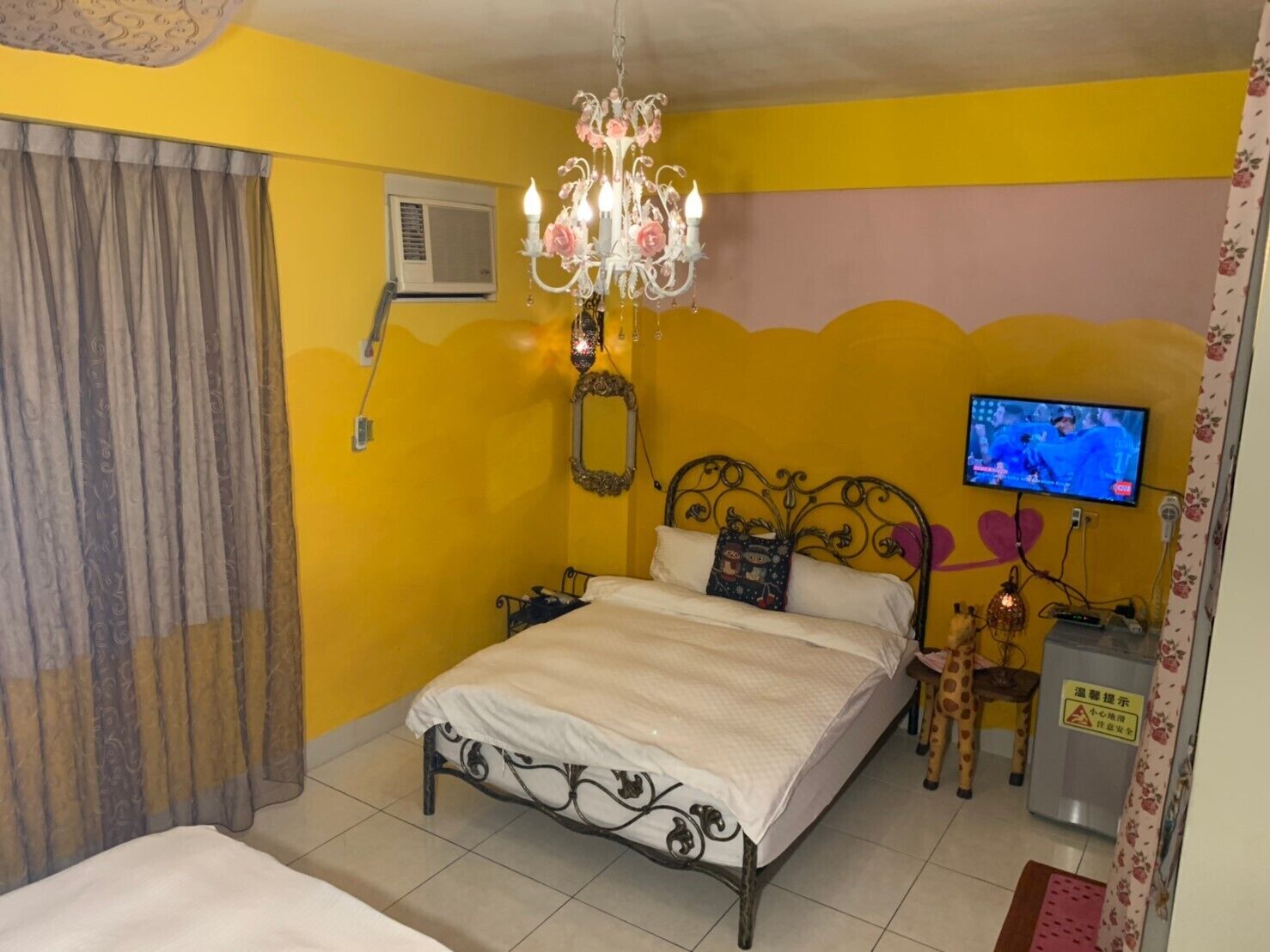 A bedroom with a bed, a chandelier, a television, and a small table with a lamp.