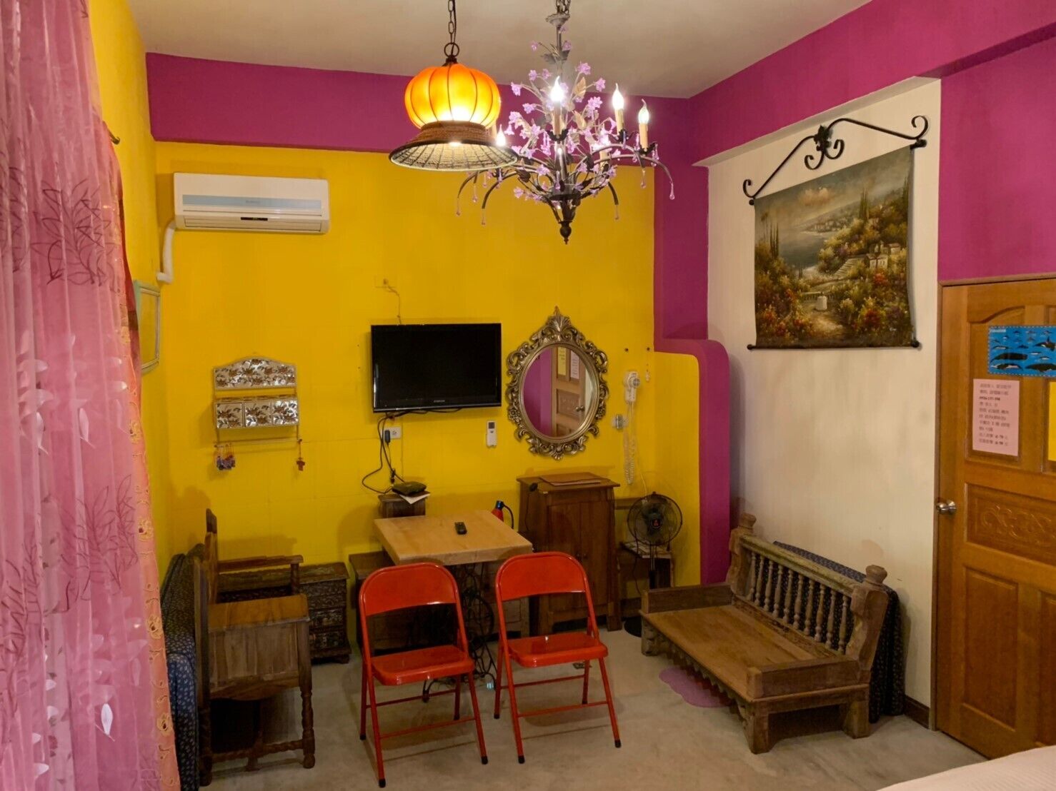 Une pièce avec un mur jaune, un banc en bois, une table avec deux chaises rouges, un lustre, une télévision et un miroir.