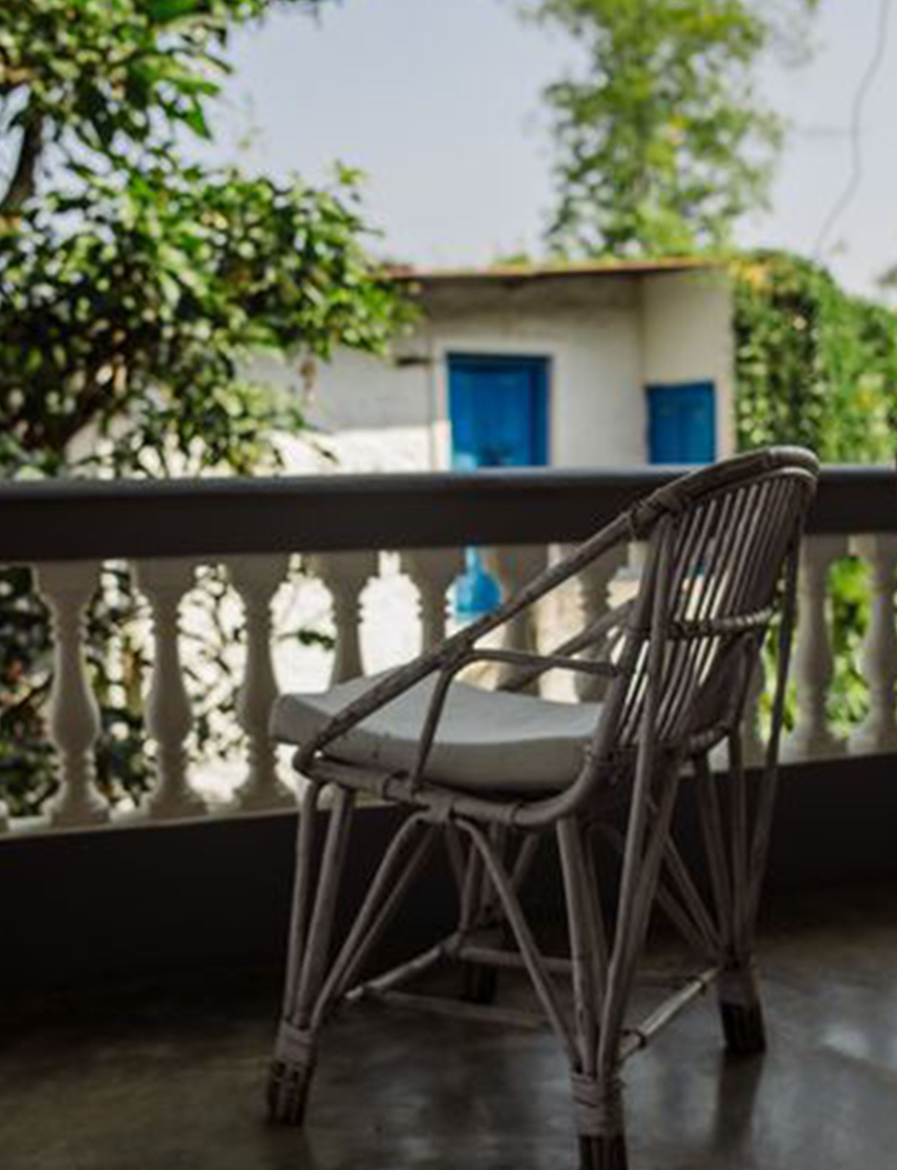Una sedia in vimini su un balcone con ringhiera, uno stabile con porte blu sullo sfondo e vegetazione tutt'intorno.