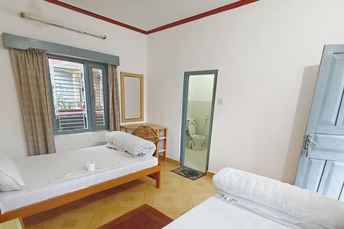 Une chambre avec deux lits, une fenêtre avec des rideaux, un ventilateur de plafond et une porte donnant sur une salle de bain.