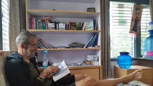 A person sitting on a couch reading a book in a room with a bookshelf, a water dispenser, and a large water container by the window. - Hotel Mountain View (Pokhara)