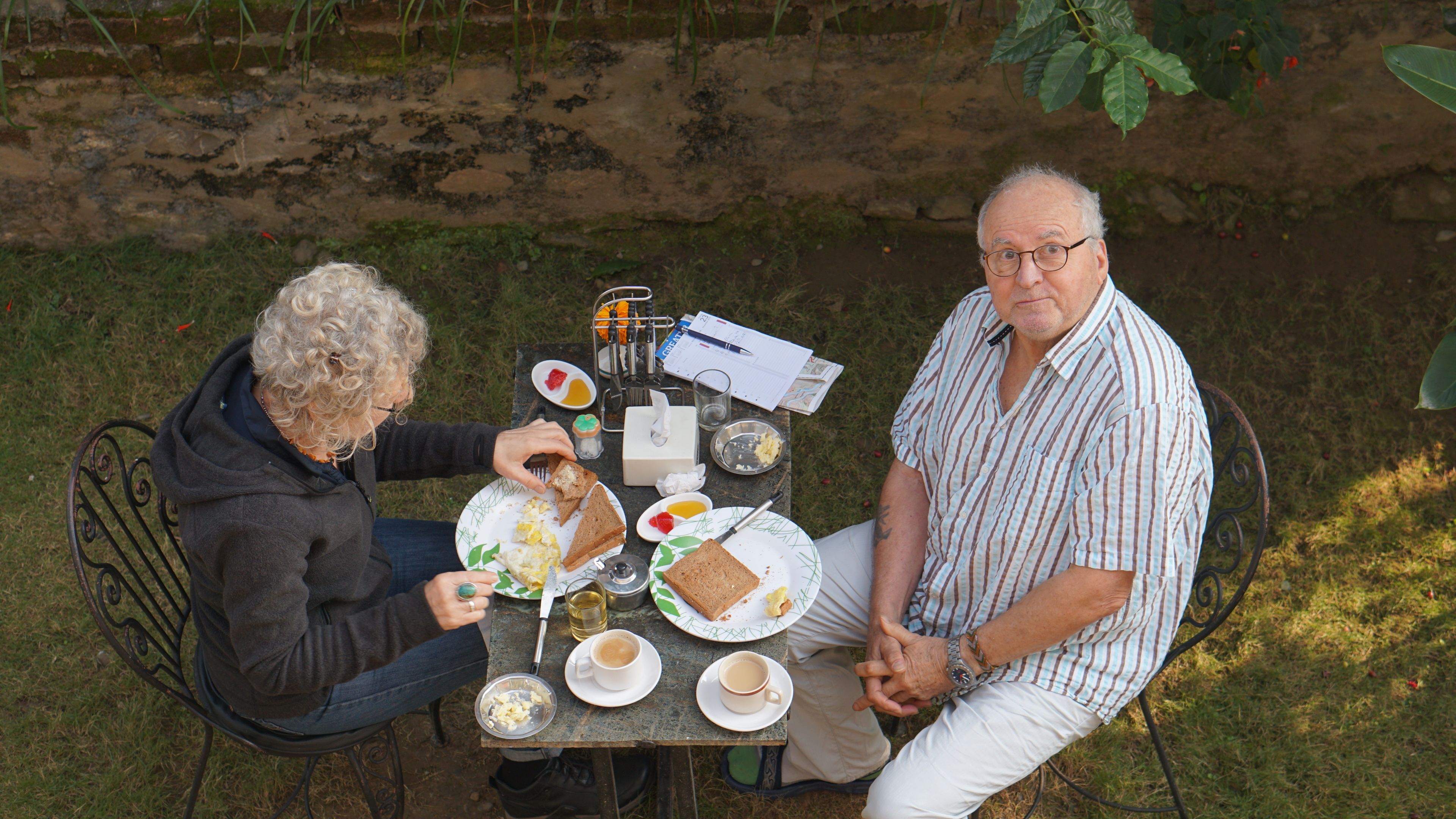 Two people seated at an outdoor table with plates of food, cups, and a notebook.