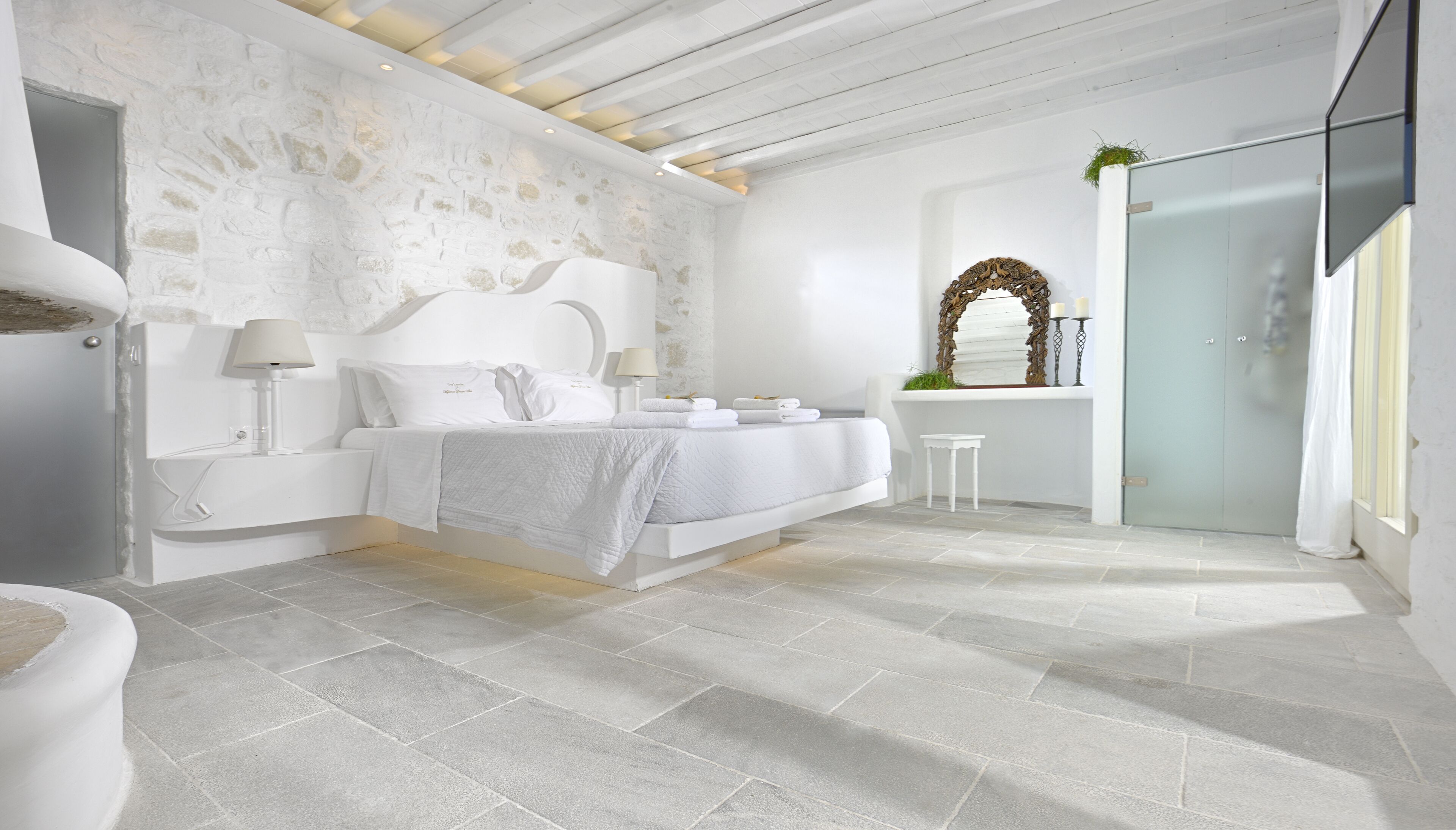 A modern bedroom with a stone wall, a bed with white linens, a bedside table with a lamp, and a small stool.