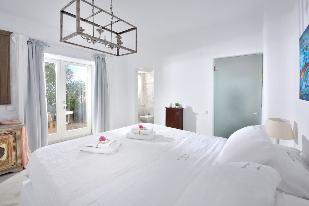 Une chambre d’hôtel avec un grand lit, une commode en bois, un bureau, une chaise, un tableau et une vue sur le jardin par la fenêtre.