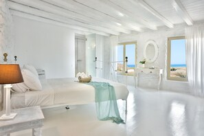 Dormitorio de tonos blancos con cama, mesita de noche y vista a la playa desde la ventana.