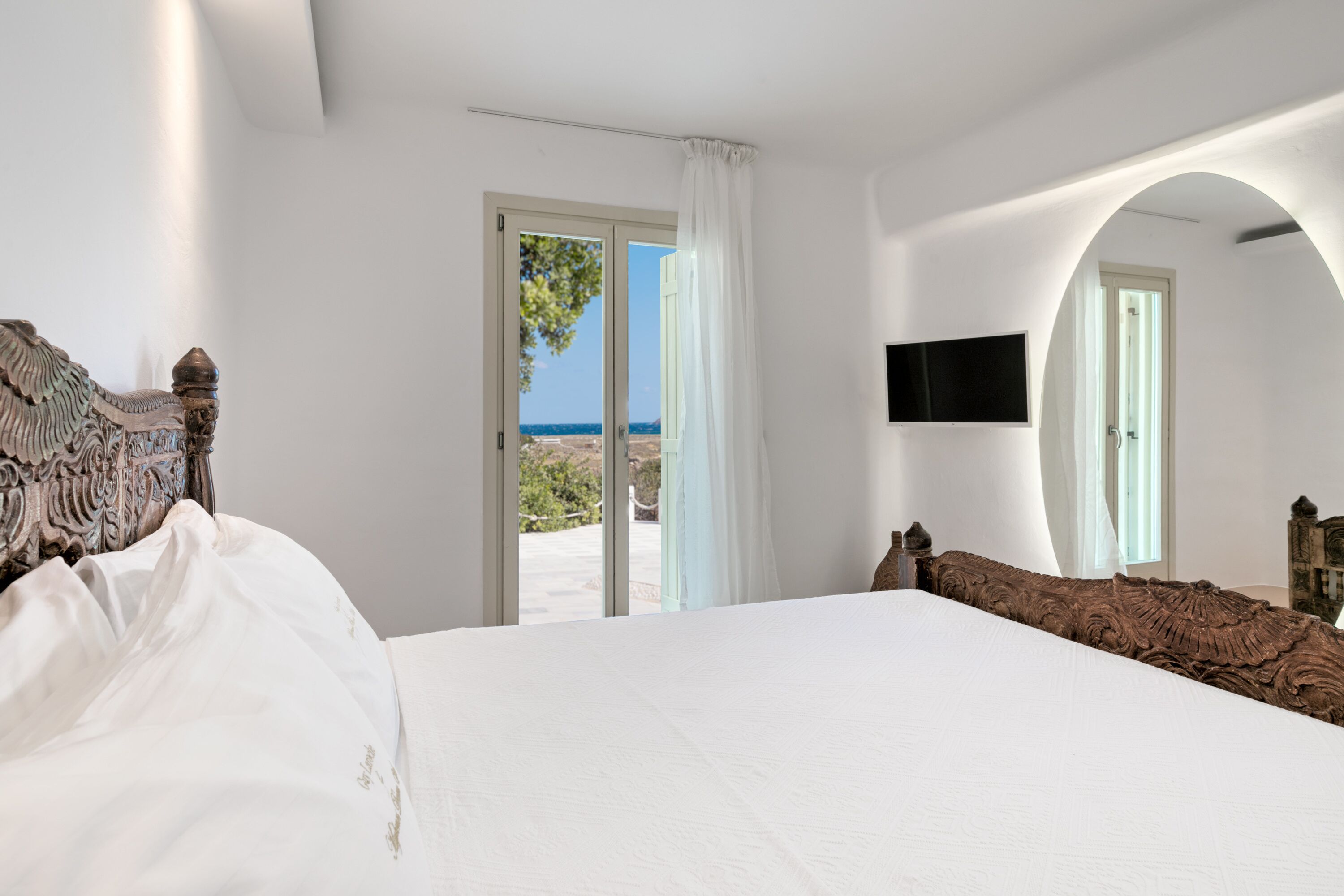 A bedroom with a large bed, a TV mounted on the wall, and a view of the outdoors through a door.