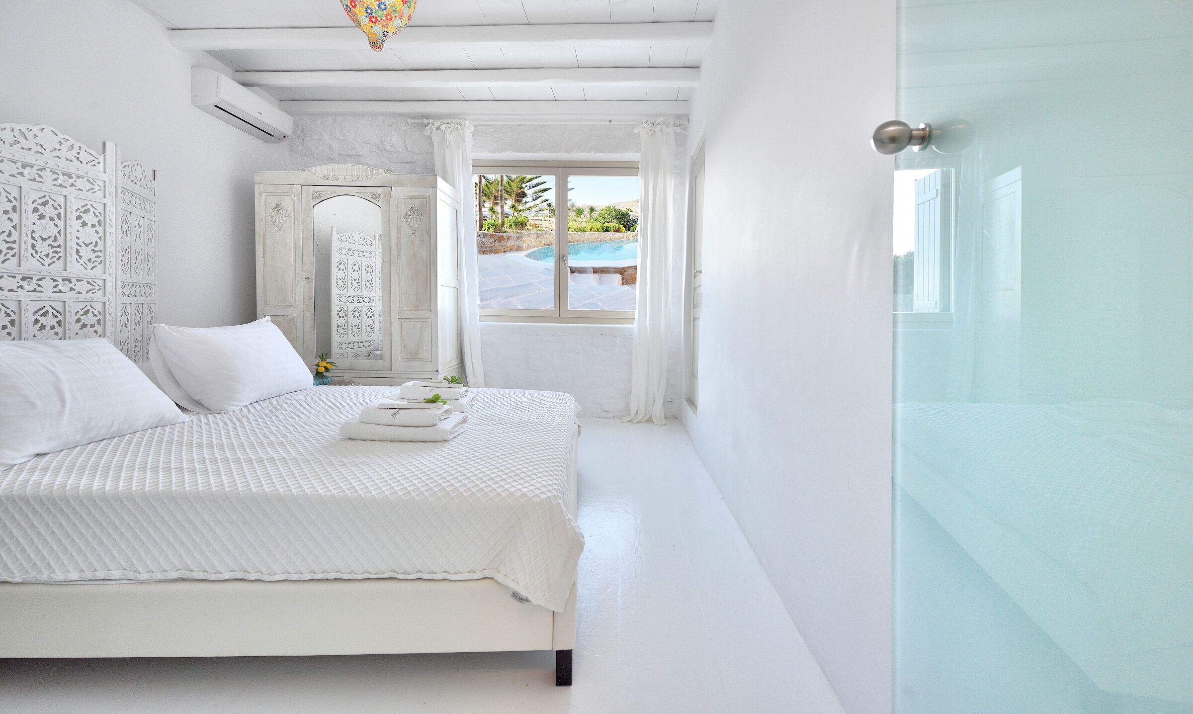 Una camera da letto bianca con un letto, uno specchio grande e vista su una piscina.