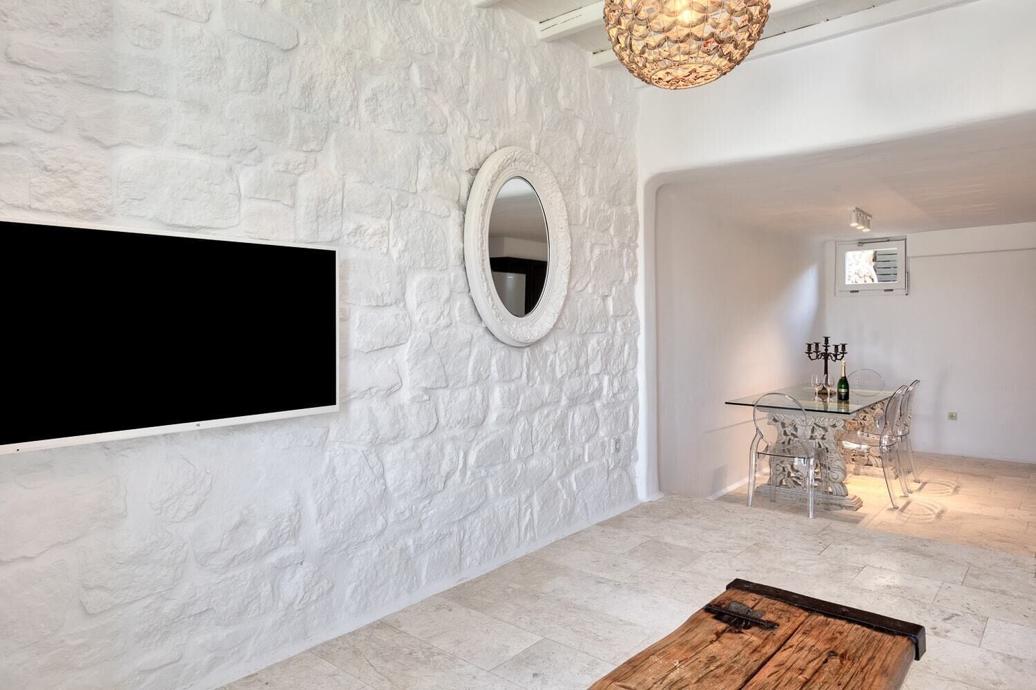Una sala moderna con pared de textura de piedra, un espejo redondo, un televisor de pantalla plana, una zona de comedor con una mesa de vidrio y sillas, y una mesa de centro de madera.