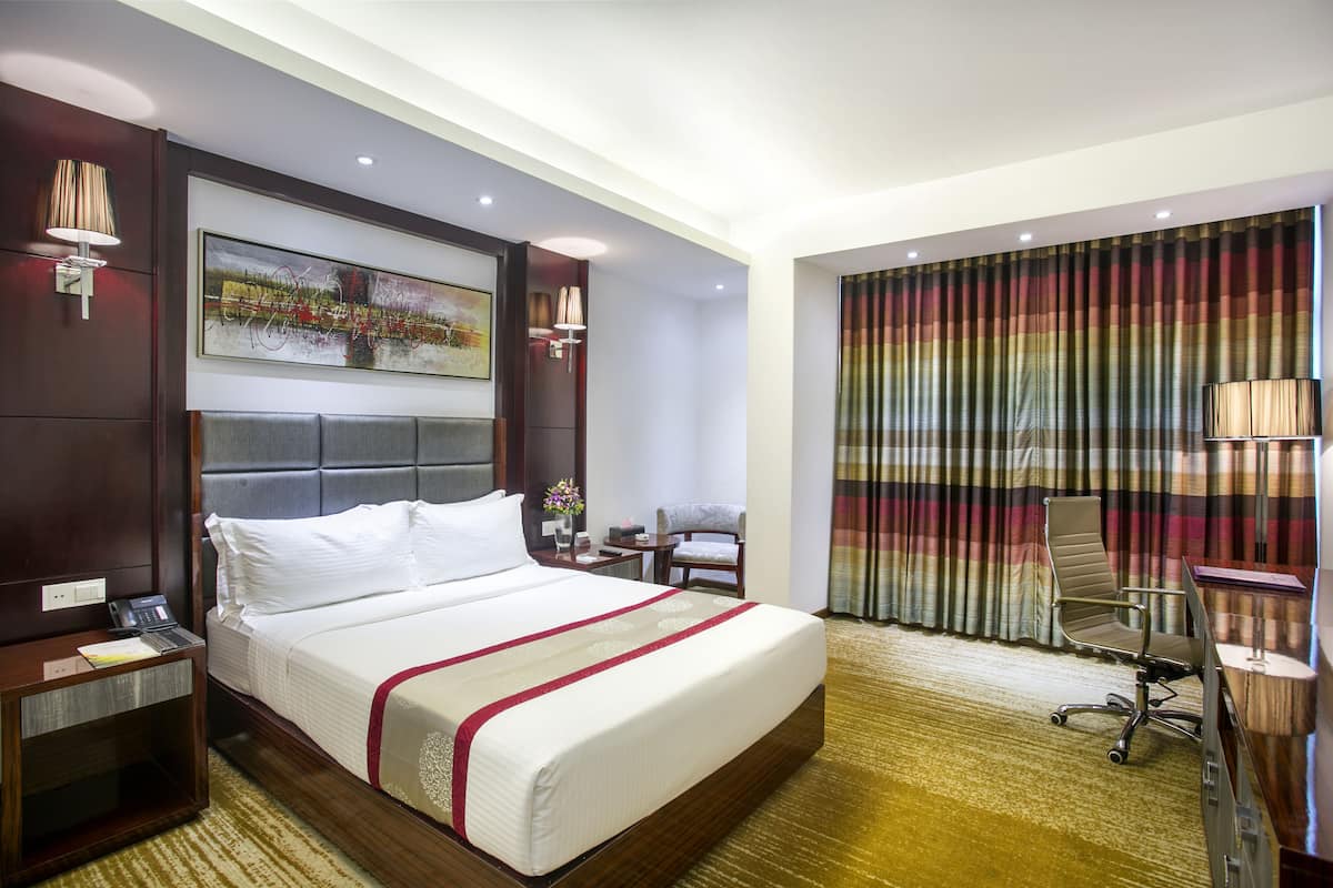 A hotel room with a large bed, a desk with a lamp, a chair, and a painting on the wall.