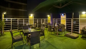 An outdoor terrace with tables and chairs, a green artificial lawn, and a view of the city at night. - Hotel Bengal Blueberry (Dhaka)