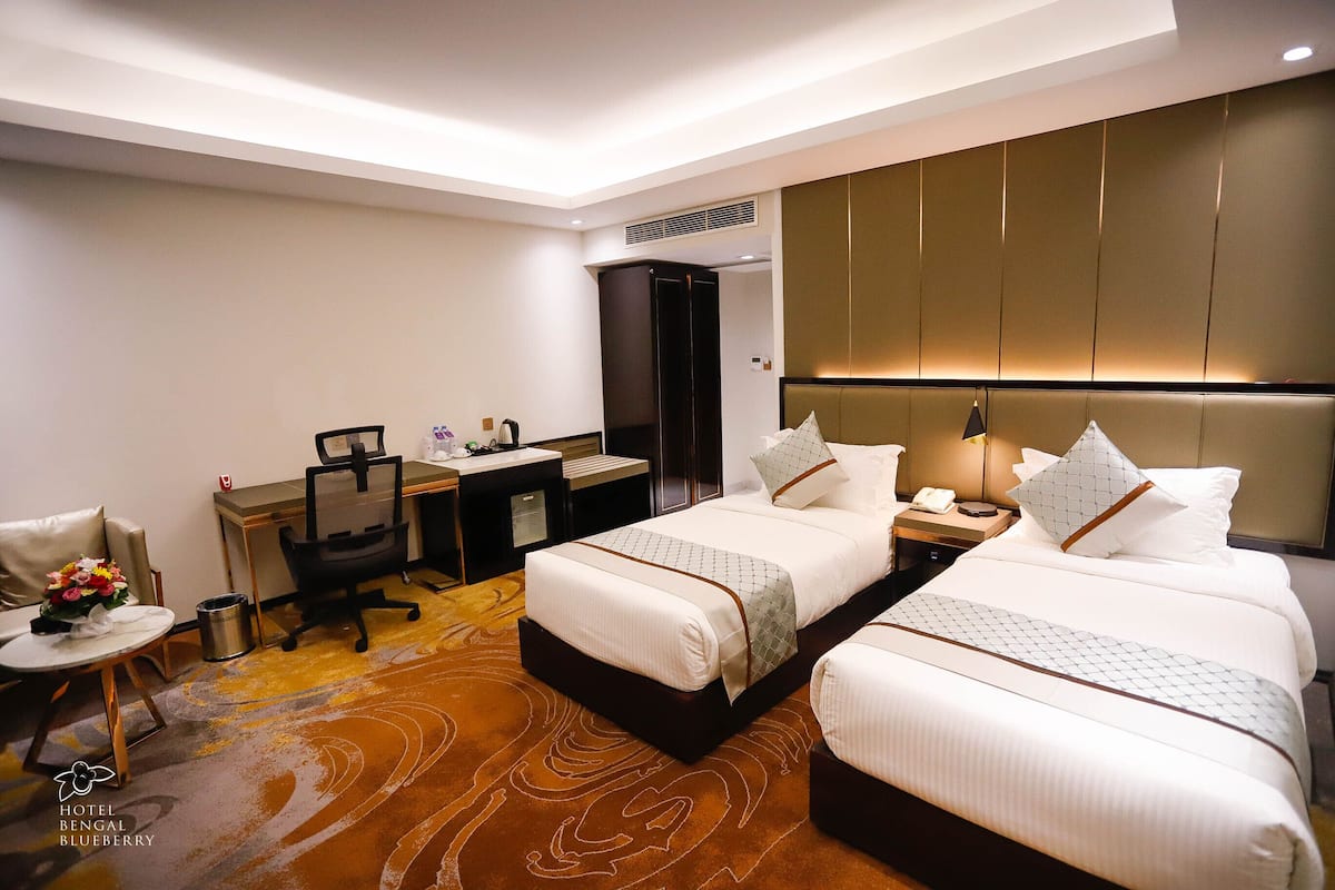 A hotel room with two beds, a desk, a chair, a small table with flowers, and a trash can.