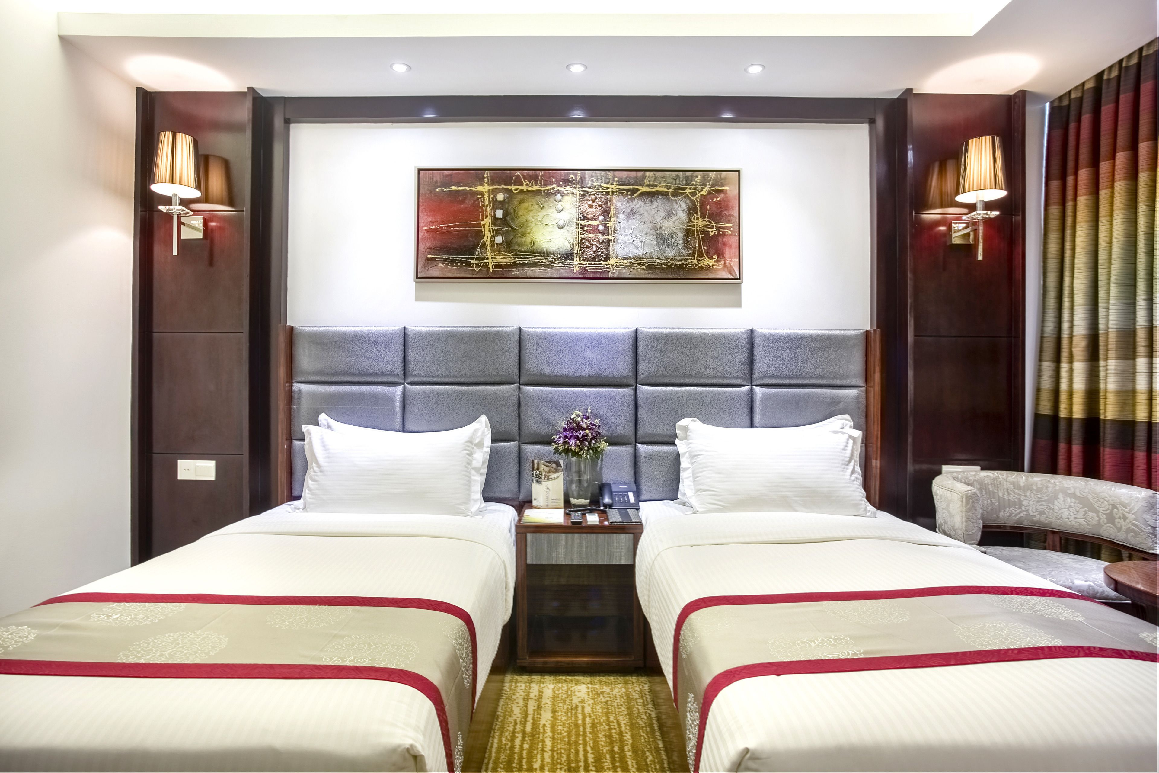 A hotel room with two beds, a large abstract painting on the wall, and a small table with a vase and a phone.