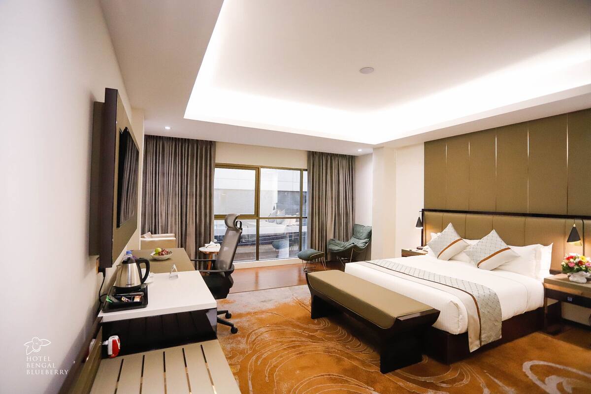 A hotel room with a bed, desk, chair, and a view of the city through the window.