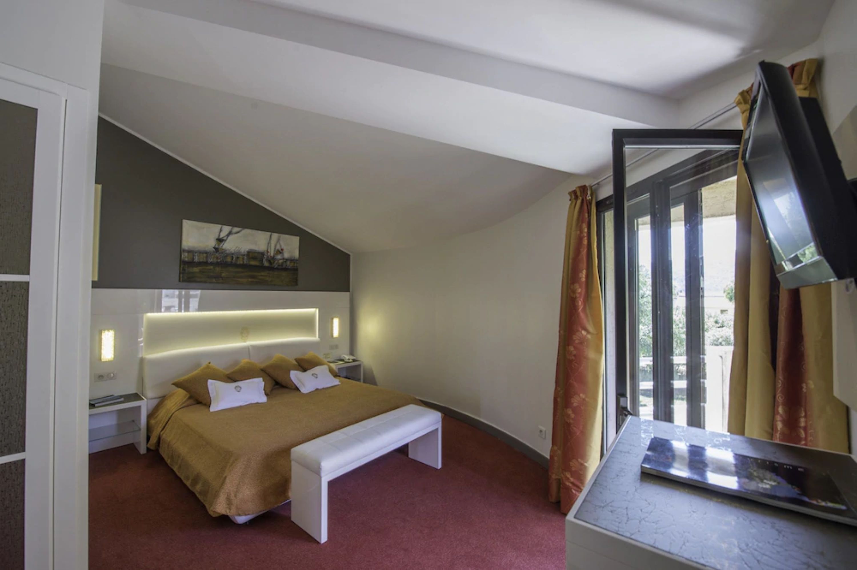 Una habitación de hotel moderna con una cama grande, un banco, un escritorio con televisor de pantalla plana y una ventana con cortinas.