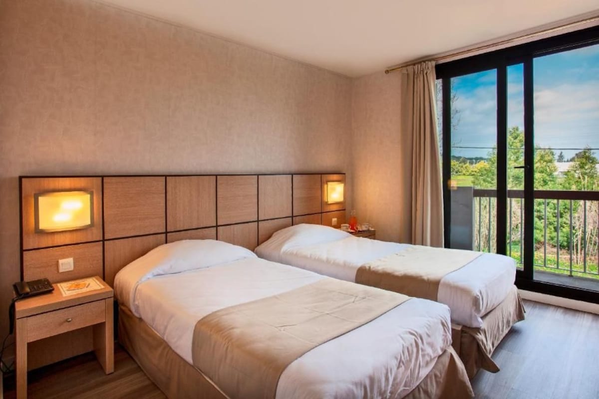 A hotel room with two beds, a nightstand, and a large window with a view of greenery.