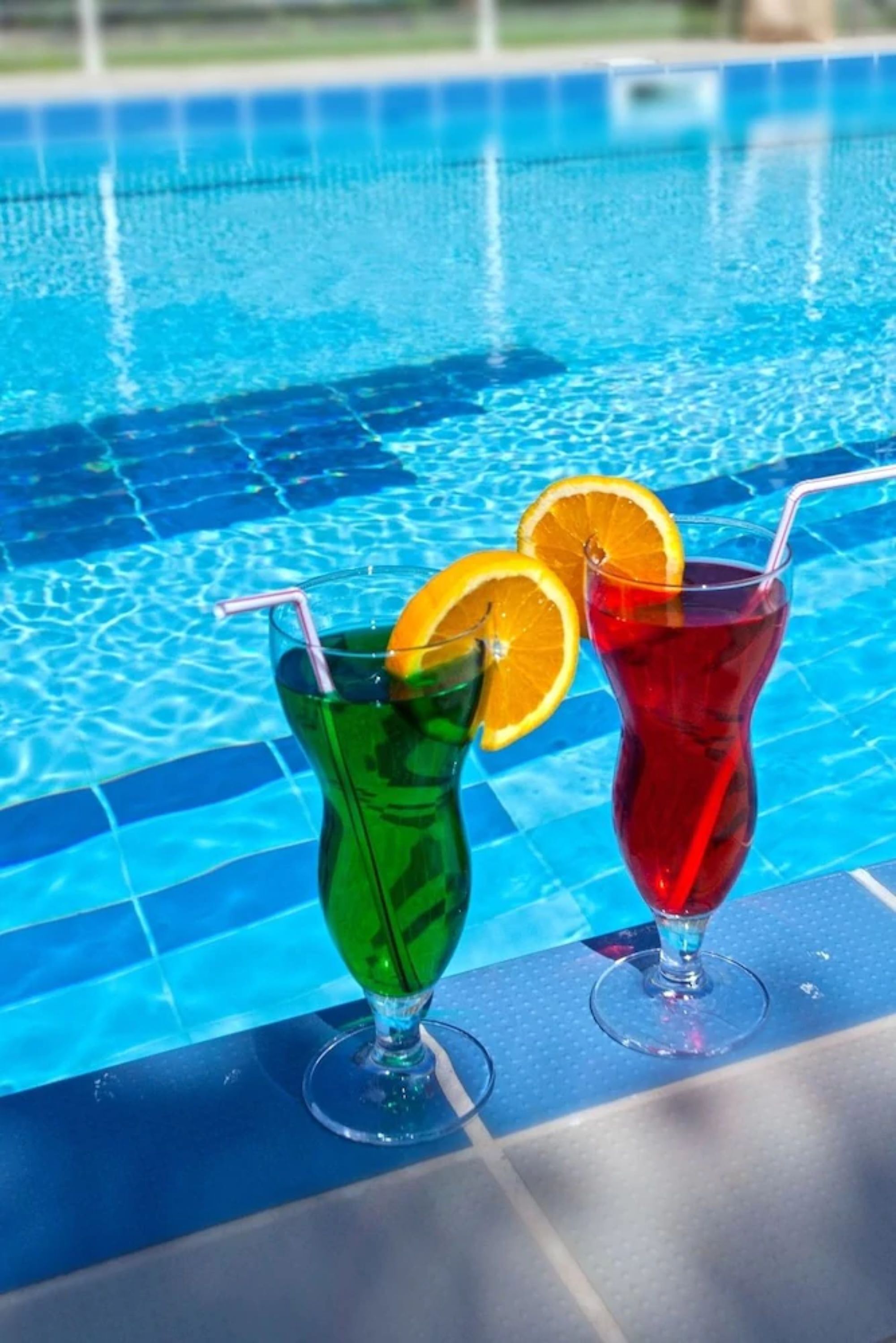 Two cocktails with straws and orange slices on a poolside table.