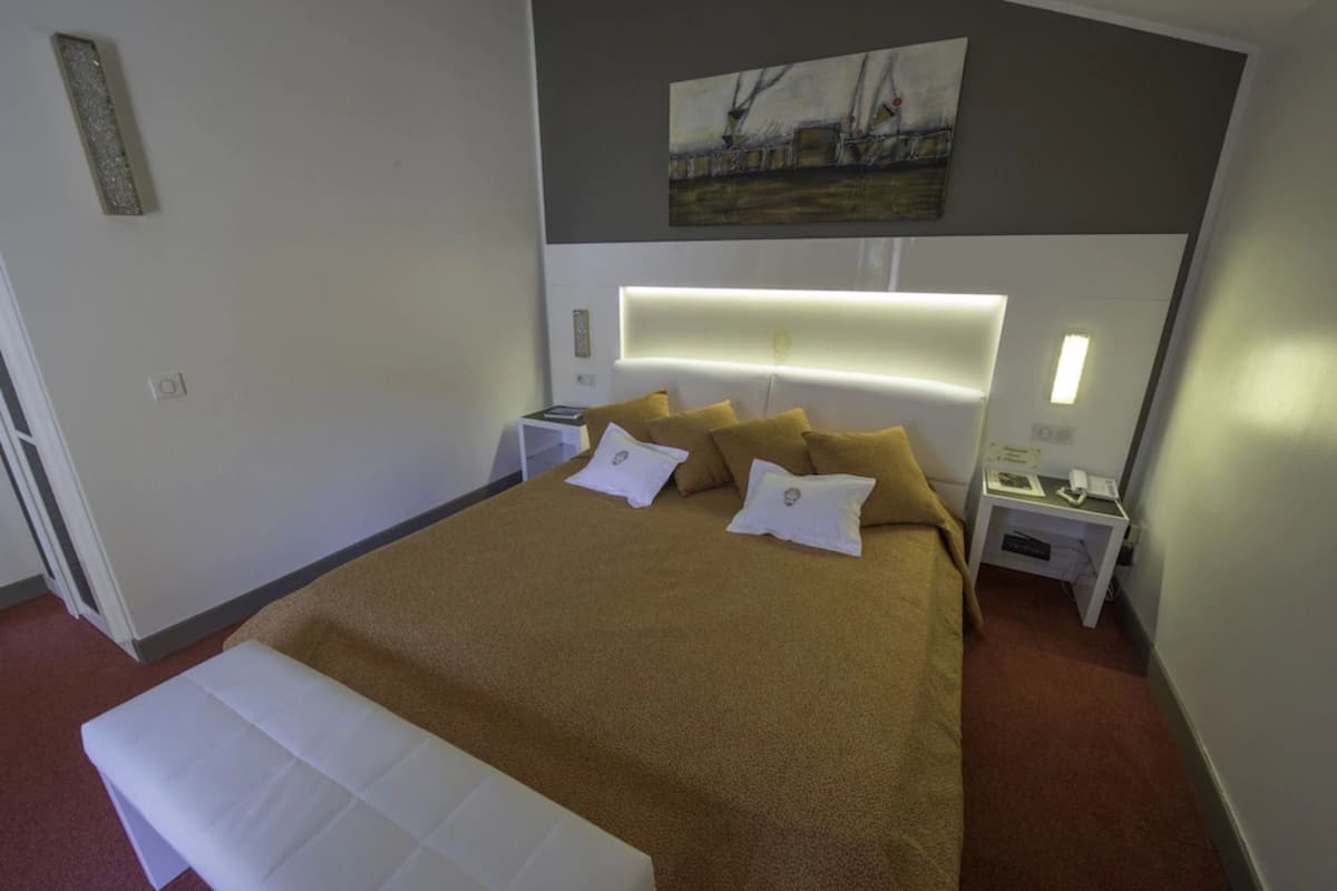 Habitación de hotel con una cama grande, cabecera con iluminación integrada, mesita de noche con un teléfono y un libro, y un cuadro en la pared.