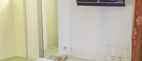 A room with a white desk, a white chair, a flat-screen TV mounted on the wall, and a mirror above the desk.