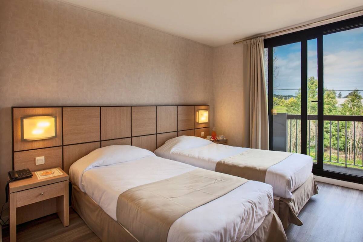 Habitación de hotel con dos camas, una mesita de noche, una lámpara y un ventanal con vistas.