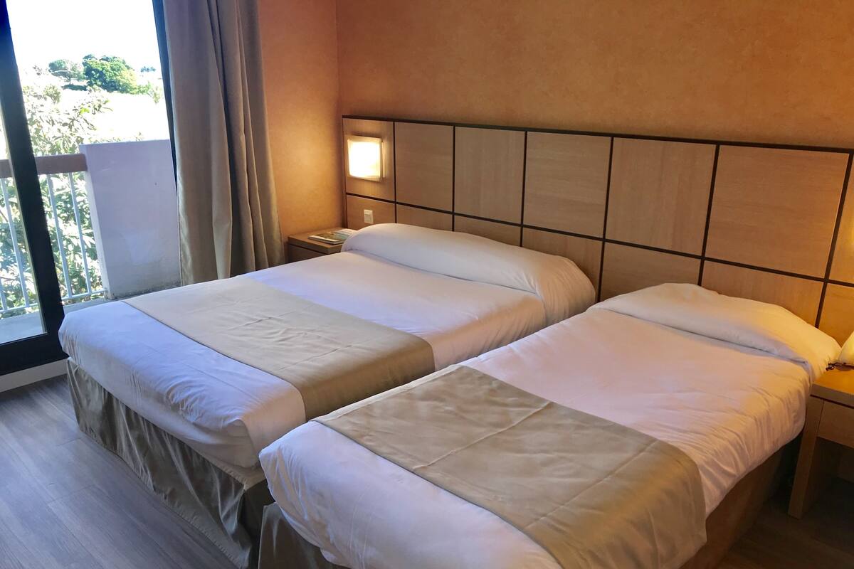 Una habitación de hotel con dos camas, una mesita de noche, una lámpara y una ventana con cortinas.