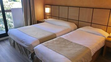 Habitació d'hotel amb dos llits, una tauleta de nit, una finestra amb cortines i vistes a vegetació.
