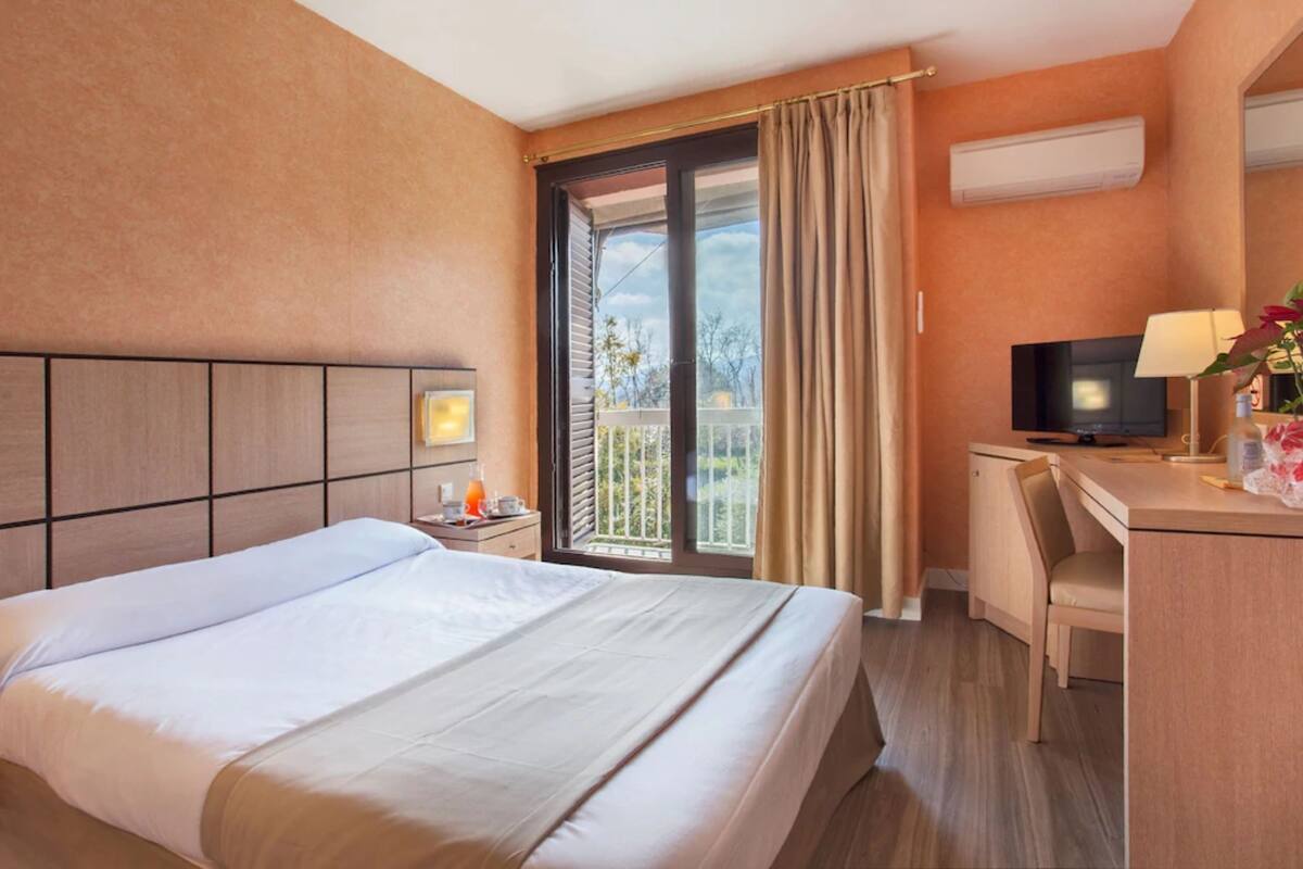 Habitación de hotel con cama, escritorio con silla, televisor y balcón con cortinas.
