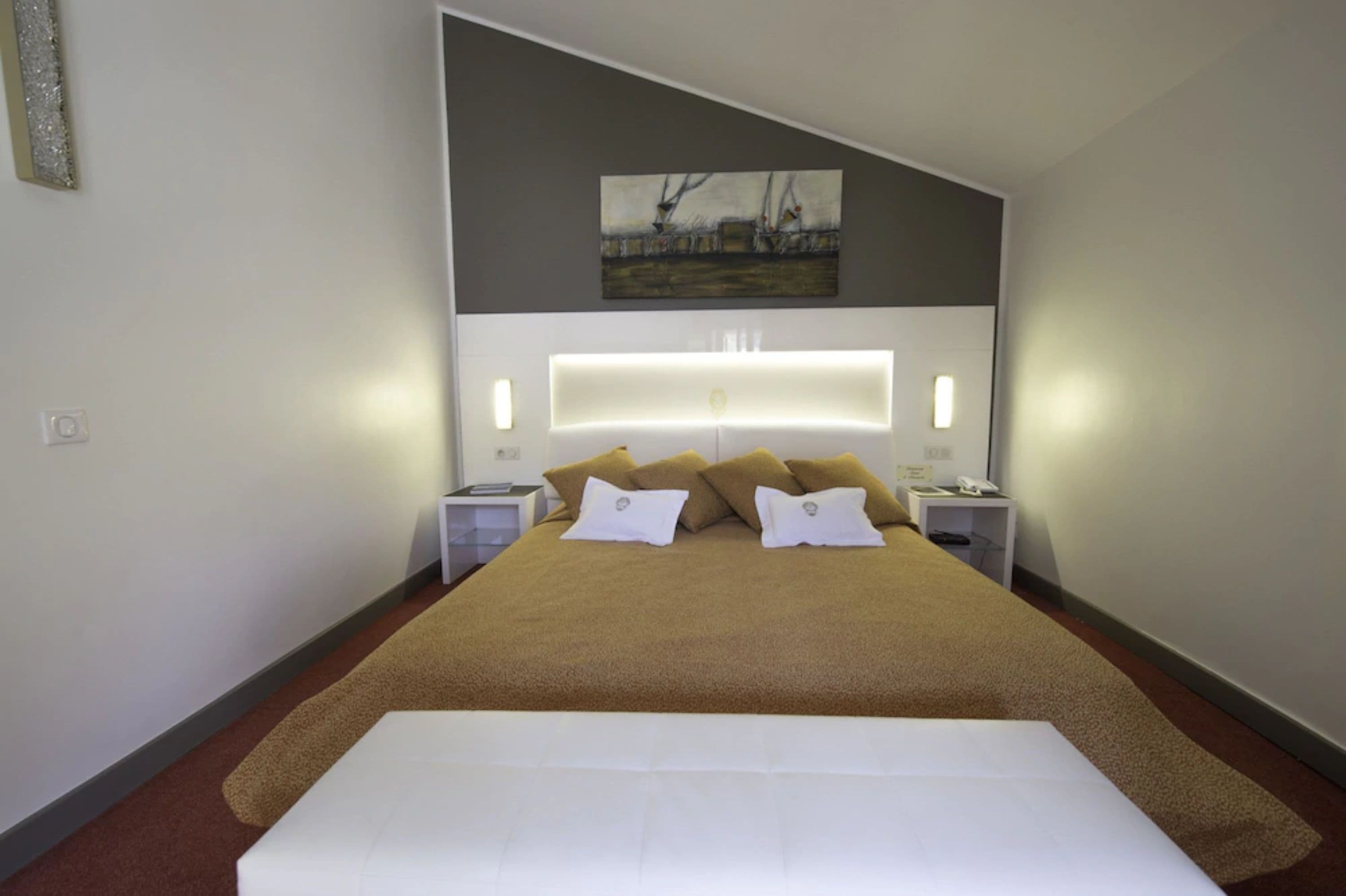 Une chambre d’hôtel moderne avec un grand lit, une tête de lit dotée d’un éclairage intégré, un tableau au mur et des tables de chevet avec des lampes.