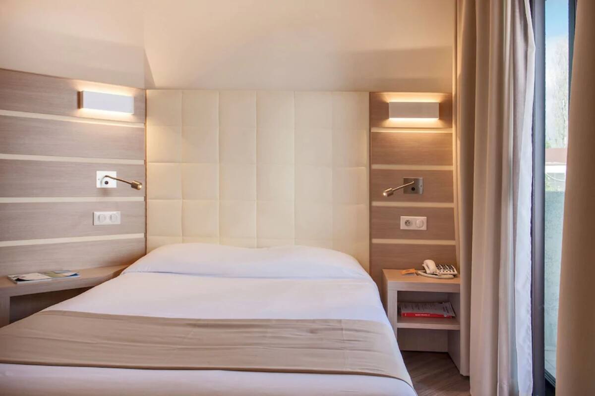 Una habitación de hotel moderna con una cama grande, mesitas de noche, una mesita auxiliar y un pequeño escritorio con un teléfono y libros.