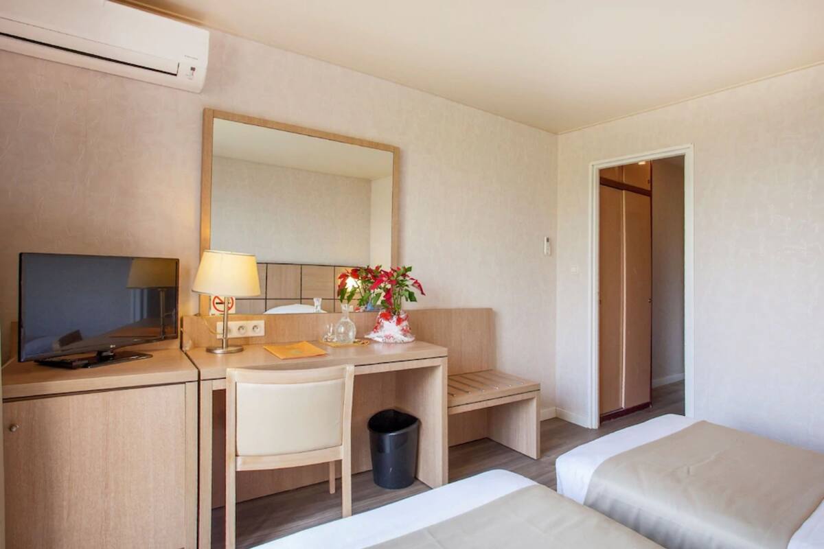 Habitación de hotel con escritorio, silla, dos camas, espejo, televisor y cesto de basura.
