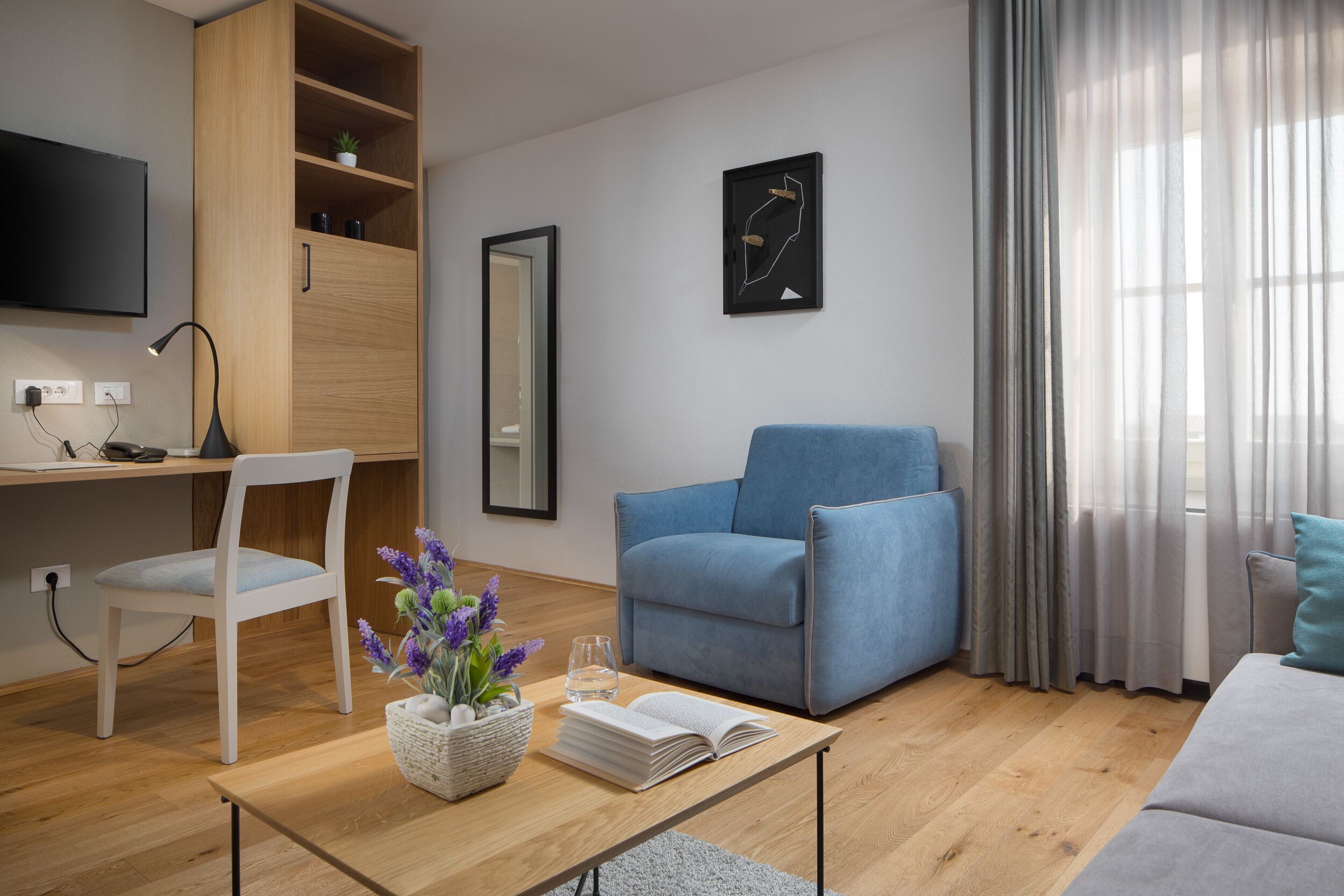 Ein modernes Wohnzimmer mit einem Holztisch, einem blauen Sessel, einem weißen Stuhl, einem Couchtisch mit Blumen in einer Vase und einem grauen Sofa.
