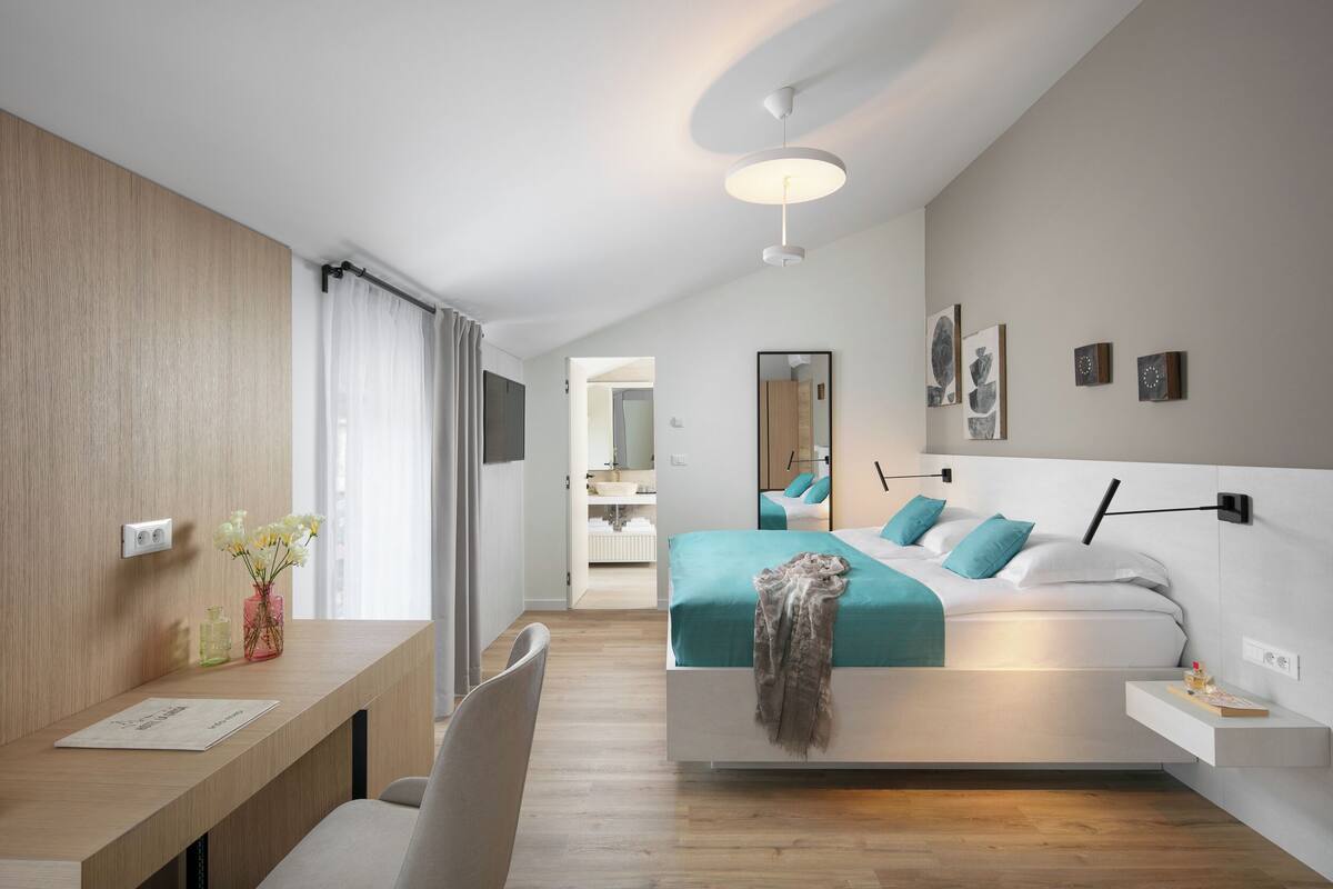 A modern hotel room with a wooden desk, a bed with teal bedding, a bedside table, and a large mirror on the wall.
