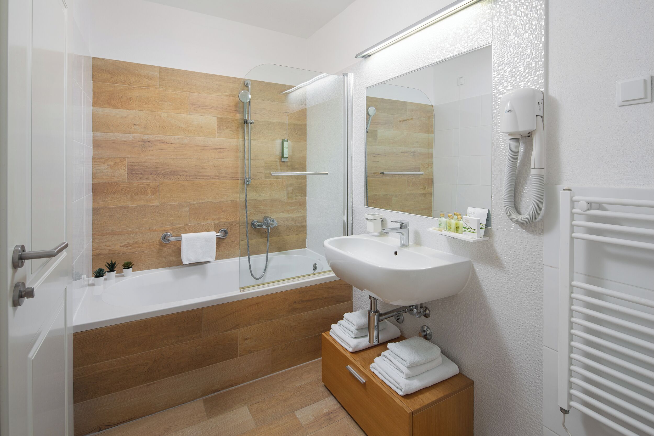 A modern bathroom with a wooden floor, a white bathtub, a sink, and a mirror.